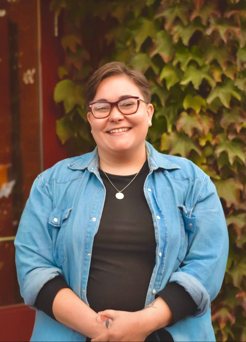 Person wearing a blue denim jacket and black shirt standing in front of a leafy green wall.